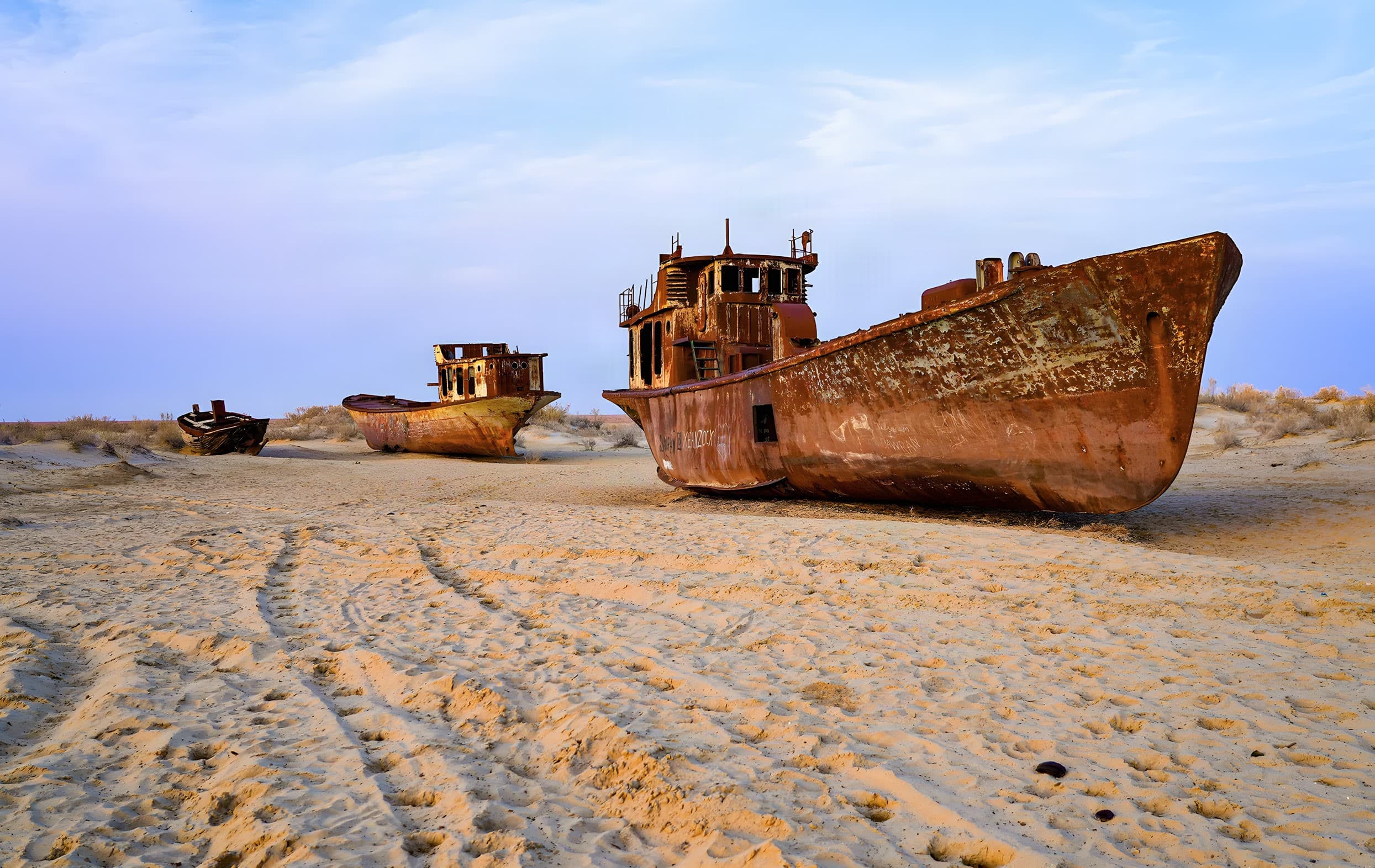 Two rusted vessels standing on sand in the area of the former port in Muynak, under a blue sky