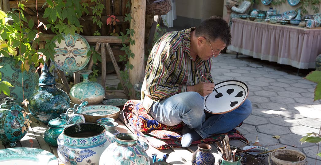 A craftsman in a skullcap sitting on the ground painting a ceramic dish, surrounded by blue and turquoise Rishtan ceramics
