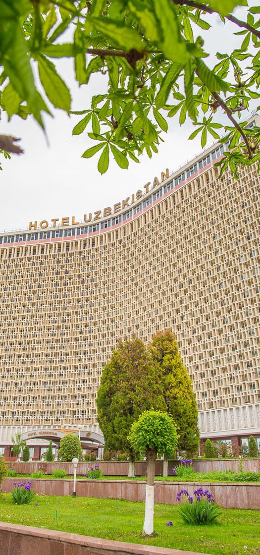 The curved façade of Hotel Uzbekistan covered in a lattice pattern, with trees and flower beds in front