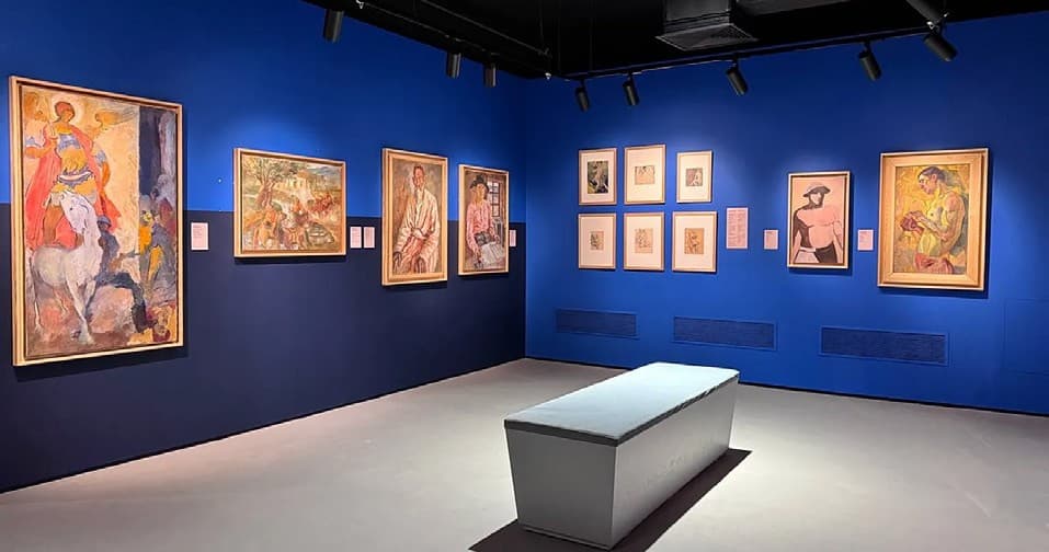 An exhibition hall with dark blue walls, bright framed paintings and a white bench in the centre