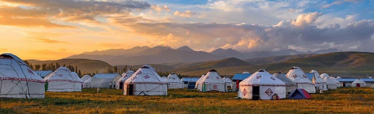 Rows of white yurts on a green meadow in a valley, with mountains behind under a sunset sky