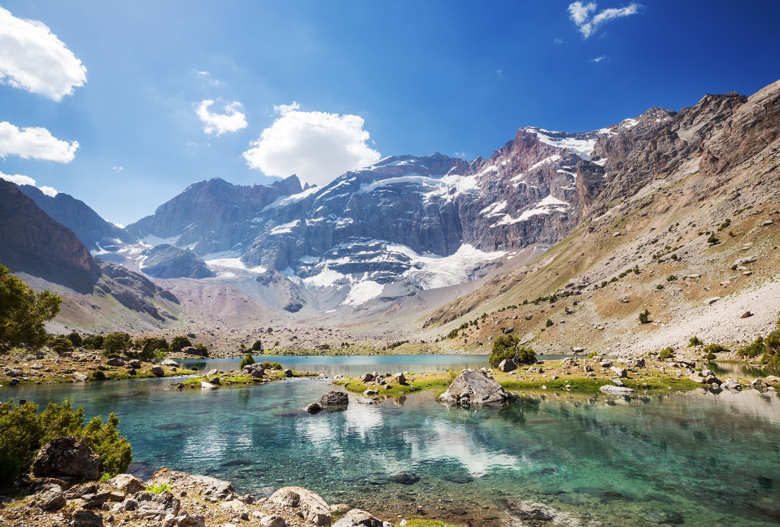 A turquoise mountain lake with boulders on the shore, beneath snow-streaked rocky peaks in the Fann Mountains
